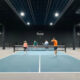 Indoor pickleball court with four players near the net; a player in orange serves as others watch, PICKLR logo on the back wall.