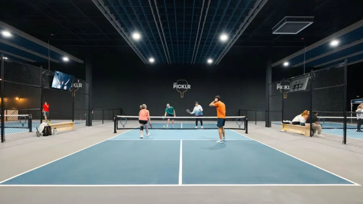 Indoor pickleball court with four players near the net; a player in orange serves as others watch, PICKLR logo on the back wall.