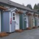 Row of colorful small cottages with white doors, wooden steps, and string lights along a gravel lot.