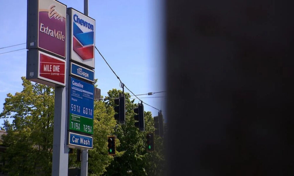 Gas station sign cluster with ExtraMile and Chevron logos, fuel-price board, and a blue sky beside green trees and a car-wash sign along a street.