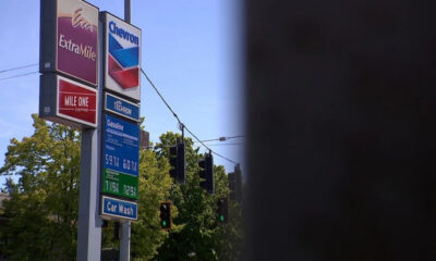 Gas station sign cluster with ExtraMile and Chevron logos, fuel-price board, and a blue sky beside green trees and a car-wash sign along a street.