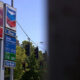 Gas station sign cluster with ExtraMile and Chevron logos, fuel-price board, and a blue sky beside green trees and a car-wash sign along a street.
