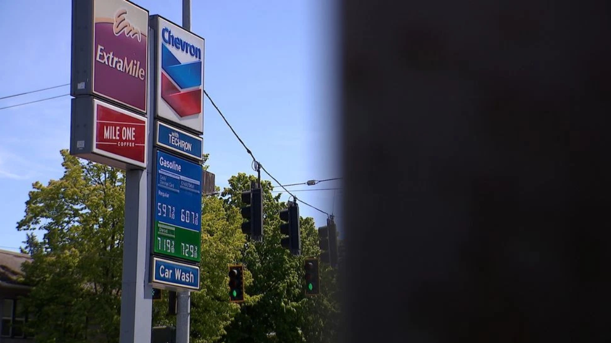 Gas station sign cluster with ExtraMile and Chevron logos, fuel-price board, and a blue sky beside green trees and a car-wash sign along a street.