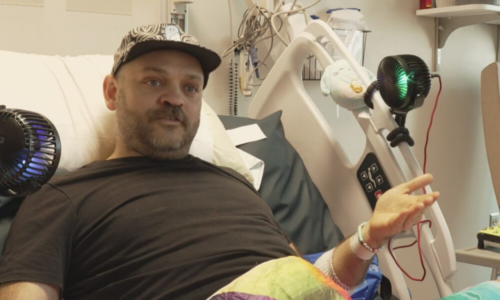 Man in a hospital bed wearing a black shirt and cap, with medical equipment and bed controls nearby.