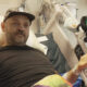 Man in a hospital bed wearing a black shirt and cap, with medical equipment and bed controls nearby.
