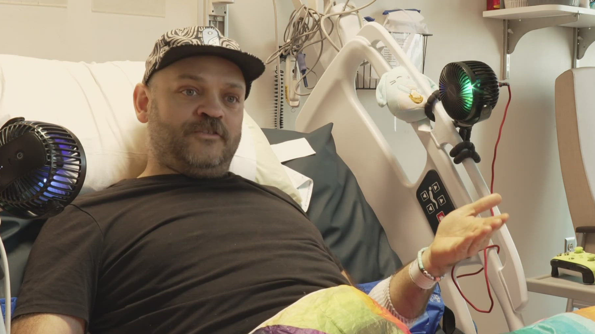 Man in a hospital bed wearing a black shirt and cap, with medical equipment and bed controls nearby.