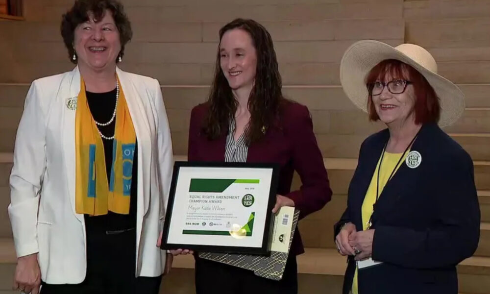 Three women stand on wooden steps; the center woman holds a framed award certificate and a gift bag, all smiling at the camera.