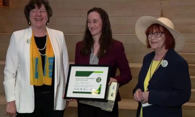Three women stand on wooden steps; the center woman holds a framed award certificate and a gift bag, all smiling at the camera.