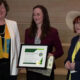 Three women stand on wooden steps; the center woman holds a framed award certificate and a gift bag, all smiling at the camera.