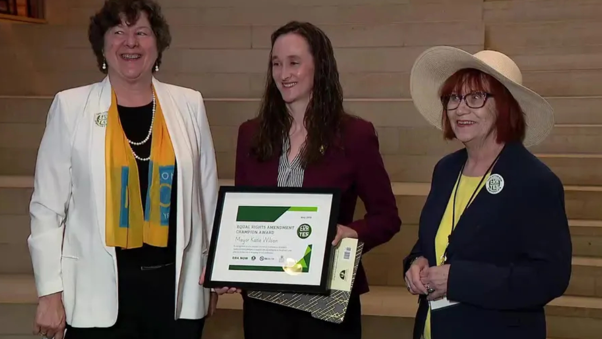 Three women stand on wooden steps; the center woman holds a framed award certificate and a gift bag, all smiling at the camera.