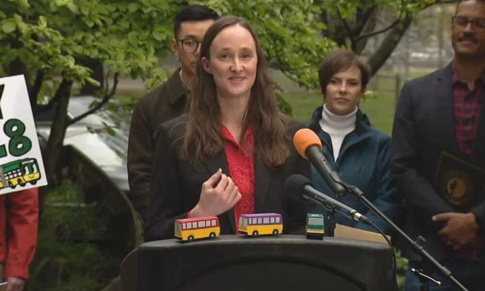 Woman in a red blouse and black blazer speaks at a podium with three colorful toy buses on the lectern, outdoor press briefing setting.