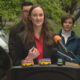 Woman in a red blouse and black blazer speaks at a podium with three colorful toy buses on the lectern, outdoor press briefing setting.