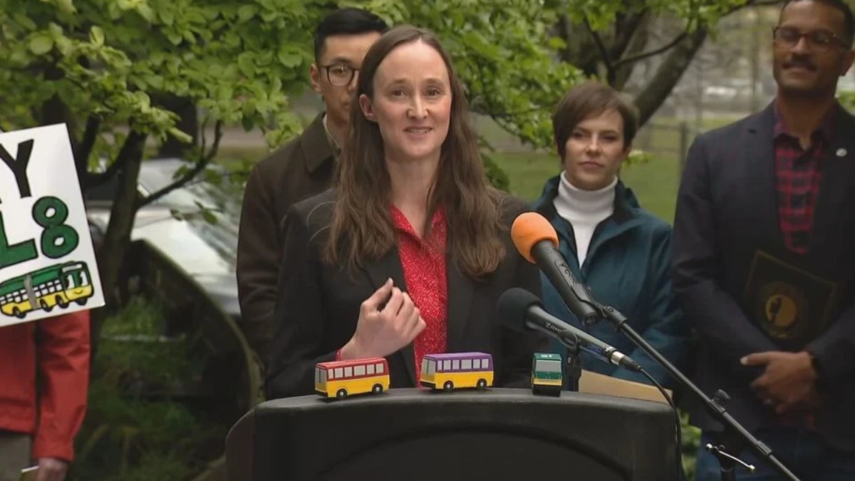 Woman in a red blouse and black blazer speaks at a podium with three colorful toy buses on the lectern, outdoor press briefing setting.