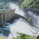 Dam releasing water over a curved spillway, with mist rising and a walkway on the left where people stand, surrounded by trees.