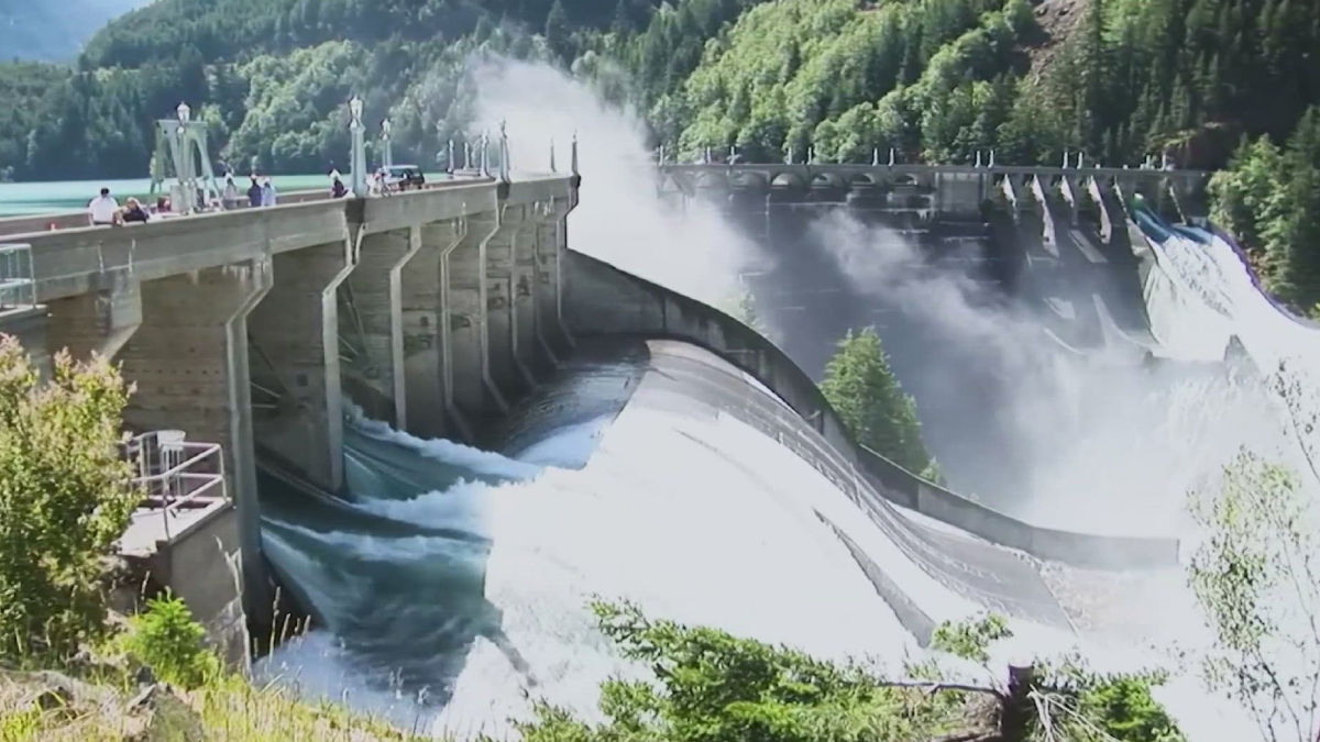 Dam releasing water over a curved spillway, with mist rising and a walkway on the left where people stand, surrounded by trees.