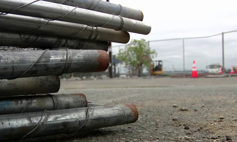 Stacked metal pipes tied together on a construction site with orange safety cones in the background.
