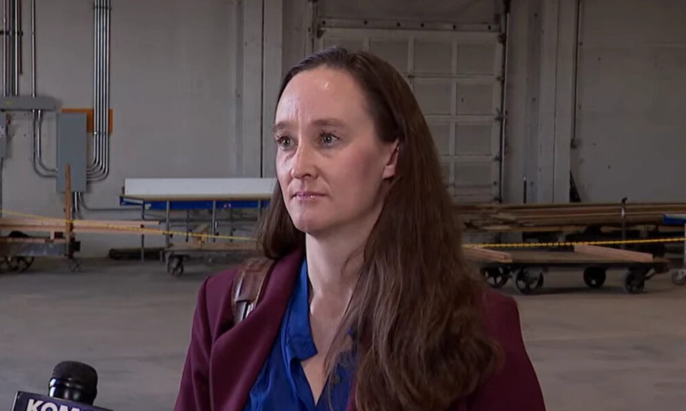 Woman in a maroon blazer being interviewed in a large industrial warehouse, with carts and pipes in the background and a microphone visible in the foreground.