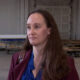Woman in a maroon blazer being interviewed in a large industrial warehouse, with carts and pipes in the background and a microphone visible in the foreground.