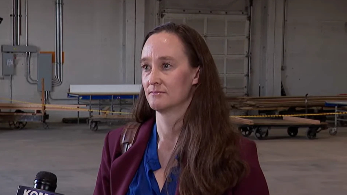 Woman in a maroon blazer being interviewed in a large industrial warehouse, with carts and pipes in the background and a microphone visible in the foreground.