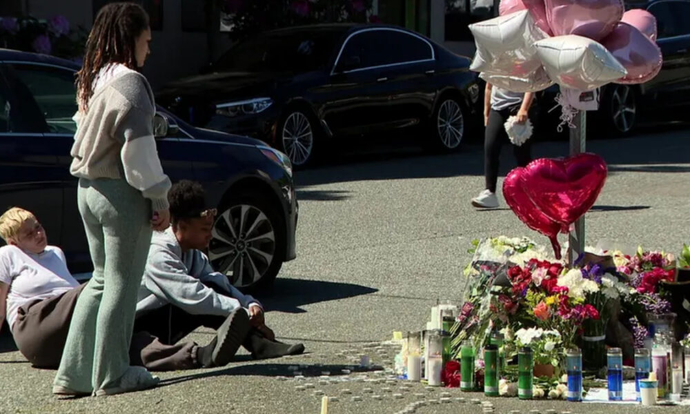 People sit on the pavement beside a roadside memorial of flowers, candles, and heart-shaped balloons beside parked cars.