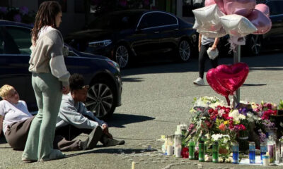 People sit on the pavement beside a roadside memorial of flowers, candles, and heart-shaped balloons beside parked cars.