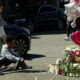People sit on the pavement beside a roadside memorial of flowers, candles, and heart-shaped balloons beside parked cars.