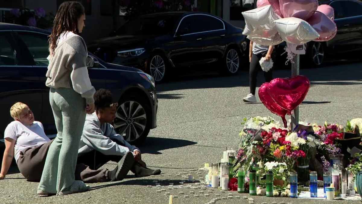 People sit on the pavement beside a roadside memorial of flowers, candles, and heart-shaped balloons beside parked cars.