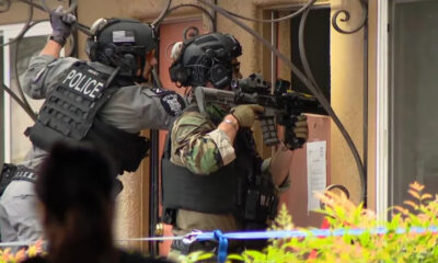 Two armed police officers in tactical gear aim rifles from a doorway, one with POLICE on his vest, preparing for a raid or standoff.