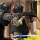 Two armed police officers in tactical gear aim rifles from a doorway, one with POLICE on his vest, preparing for a raid or standoff.