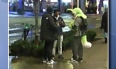 Three people stand on a wet city sidewalk, two in dark coats and sneakers, one in a bright neon jacket, chatting near a railing.