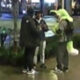 Three people stand on a wet city sidewalk, two in dark coats and sneakers, one in a bright neon jacket, chatting near a railing.