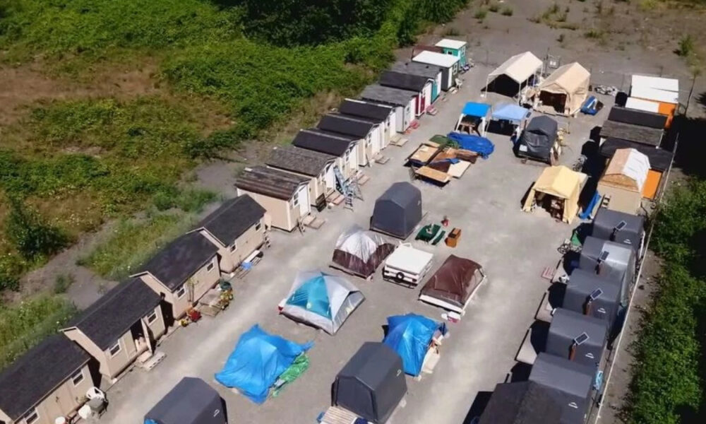 Aerial view of a temporary shelter camp with rows of small beige modular houses along the edges and tents and canopies in the central yard.