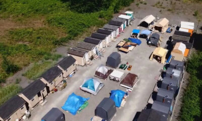 Aerial view of a temporary shelter camp with rows of small beige modular houses along the edges and tents and canopies in the central yard.