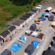 Aerial view of a temporary shelter camp with rows of small beige modular houses along the edges and tents and canopies in the central yard.