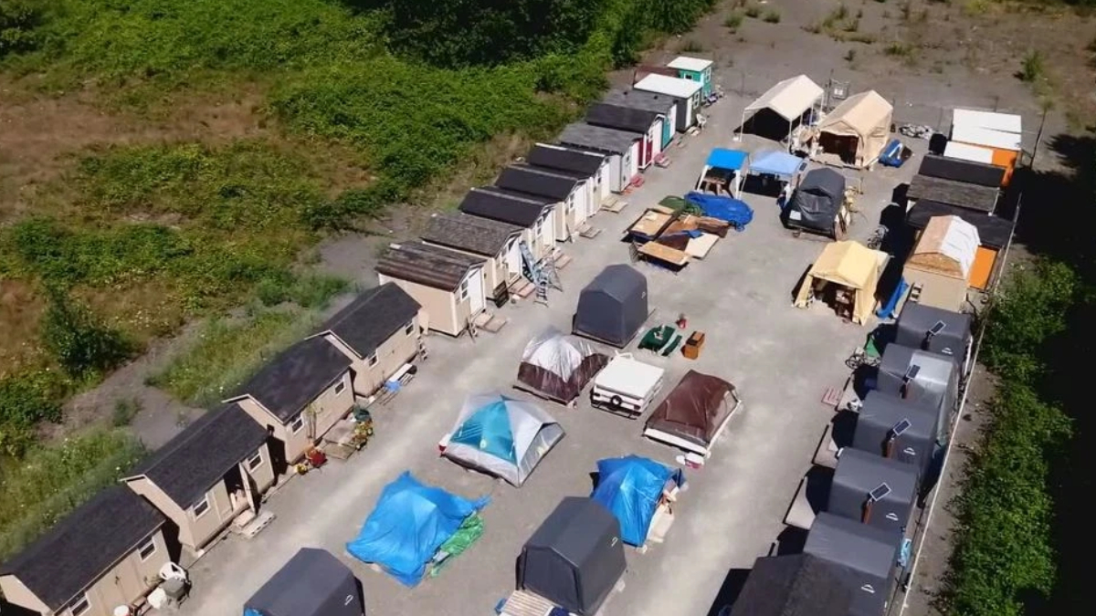 Aerial view of a temporary shelter camp with rows of small beige modular houses along the edges and tents and canopies in the central yard.