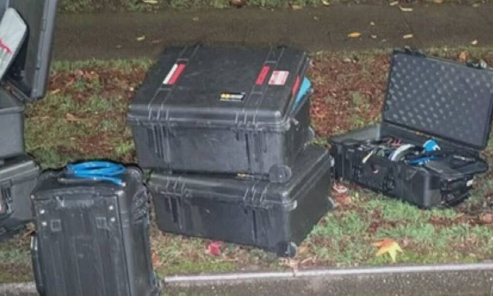 Assorted black hard protective cases on a grassy roadside with cables and tools spilling out nearby.