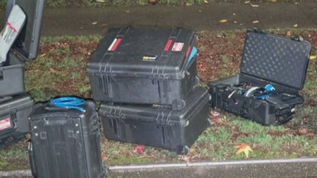 Assorted black hard protective cases on a grassy roadside with cables and tools spilling out nearby.