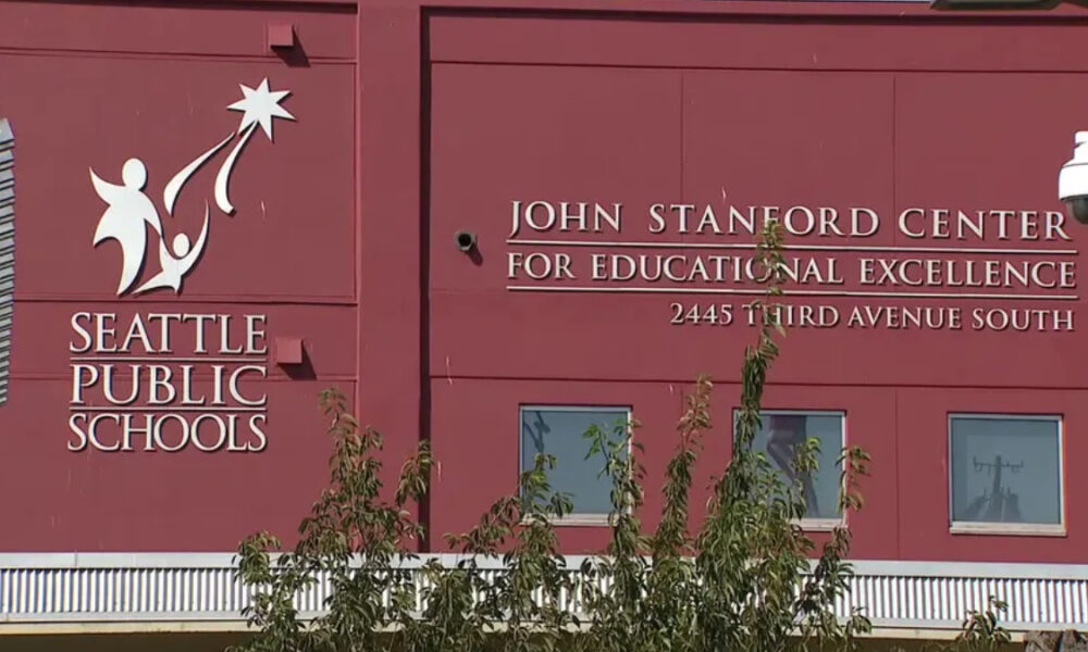 Exterior of a red Seattle Public Schools building featuring the John Stanford Center for Educational Excellence sign and the address 2445 Third Avenue South, with a tree partially obscuring the lower facade.