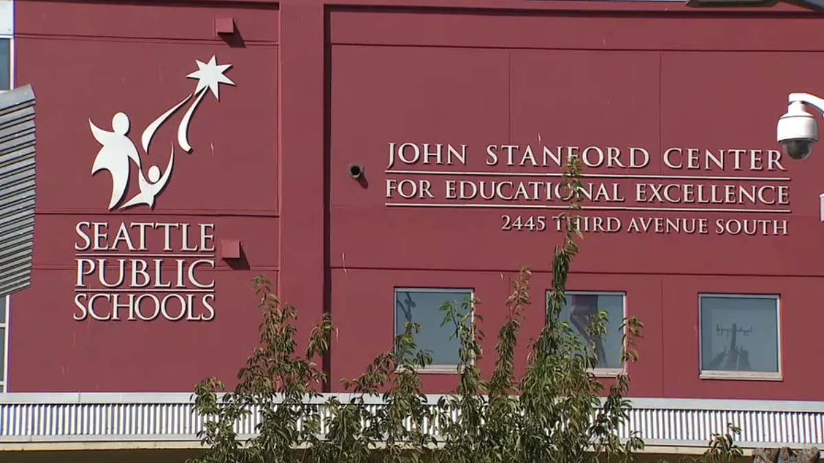 Exterior of a red Seattle Public Schools building featuring the John Stanford Center for Educational Excellence sign and the address 2445 Third Avenue South, with a tree partially obscuring the lower facade.