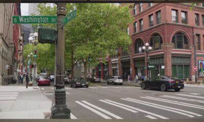 Intersection at S Washington St and 1st Ave S in a city, brick buildings, trees, crosswalks, and moving cars.