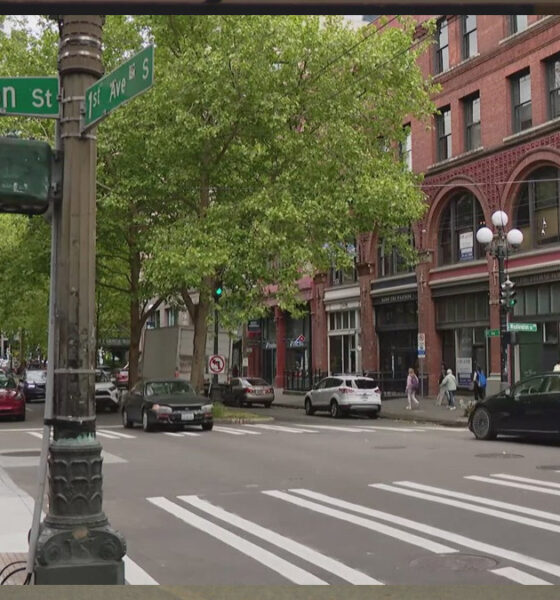 Intersection at S Washington St and 1st Ave S in a city, brick buildings, trees, crosswalks, and moving cars.