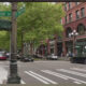 Intersection at S Washington St and 1st Ave S in a city, brick buildings, trees, crosswalks, and moving cars.