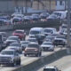 Heavy traffic on a multi-lane highway with concrete barriers and orange construction cones along the median edge.
