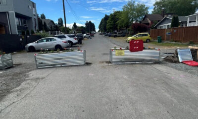 A residential street closed off by three metal barriers with a yellow warning sign and red safety cage, cars parked along both sides, and construction debris on the right.