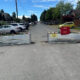 A residential street closed off by three metal barriers with a yellow warning sign and red safety cage, cars parked along both sides, and construction debris on the right.
