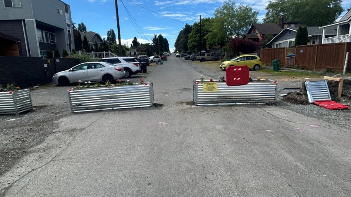 A residential street closed off by three metal barriers with a yellow warning sign and red safety cage, cars parked along both sides, and construction debris on the right.