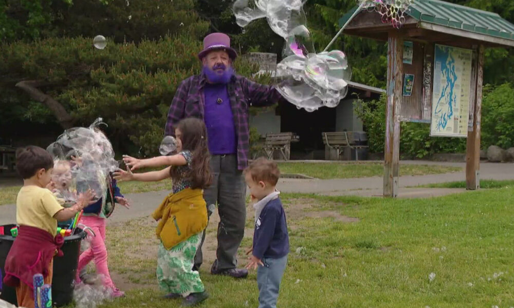 Bubble artist in a purple hat and shirt blows giant bubbles for kids in a park.