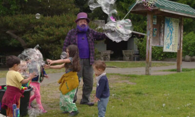 Bubble artist in a purple hat and shirt blows giant bubbles for kids in a park.
