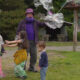 Bubble artist in a purple hat and shirt blows giant bubbles for kids in a park.