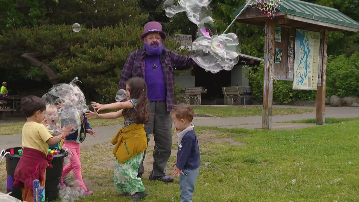 Bubble artist in a purple hat and shirt blows giant bubbles for kids in a park.